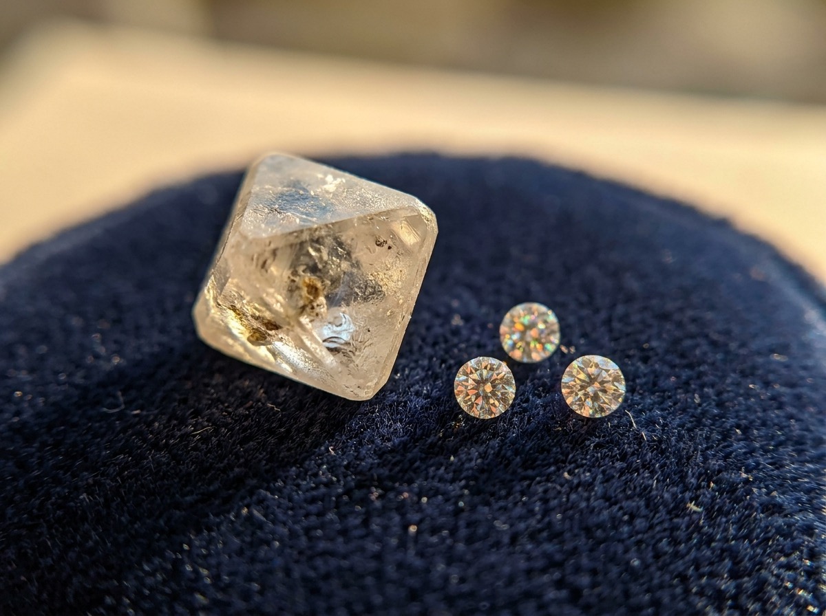 A small rough diamond crystal beside several finished round brilliant melee diamonds on navy velvet