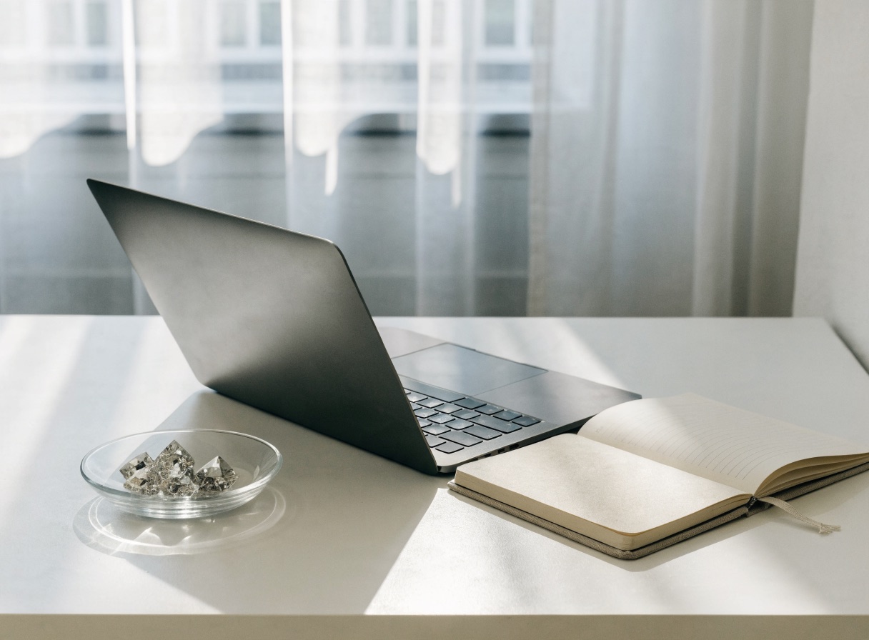 An open laptop and notebook on a clean white desk with a small glass dish holding a few loose diamonds
